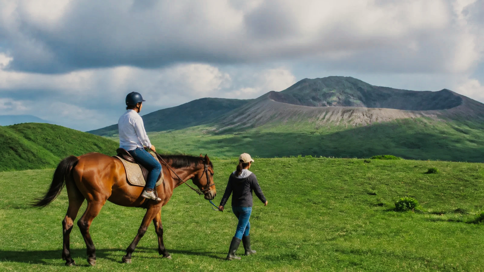 阿蘇を思わせる草原でスタッフに引かれた馬に乗る旅行者