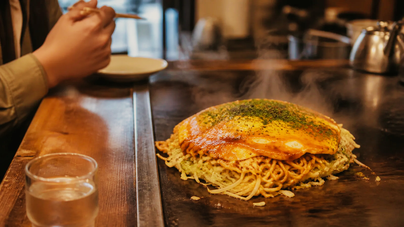 広島駅の鉄板カウンターで麺入りお好み焼きを待つ場面の編集イメージ